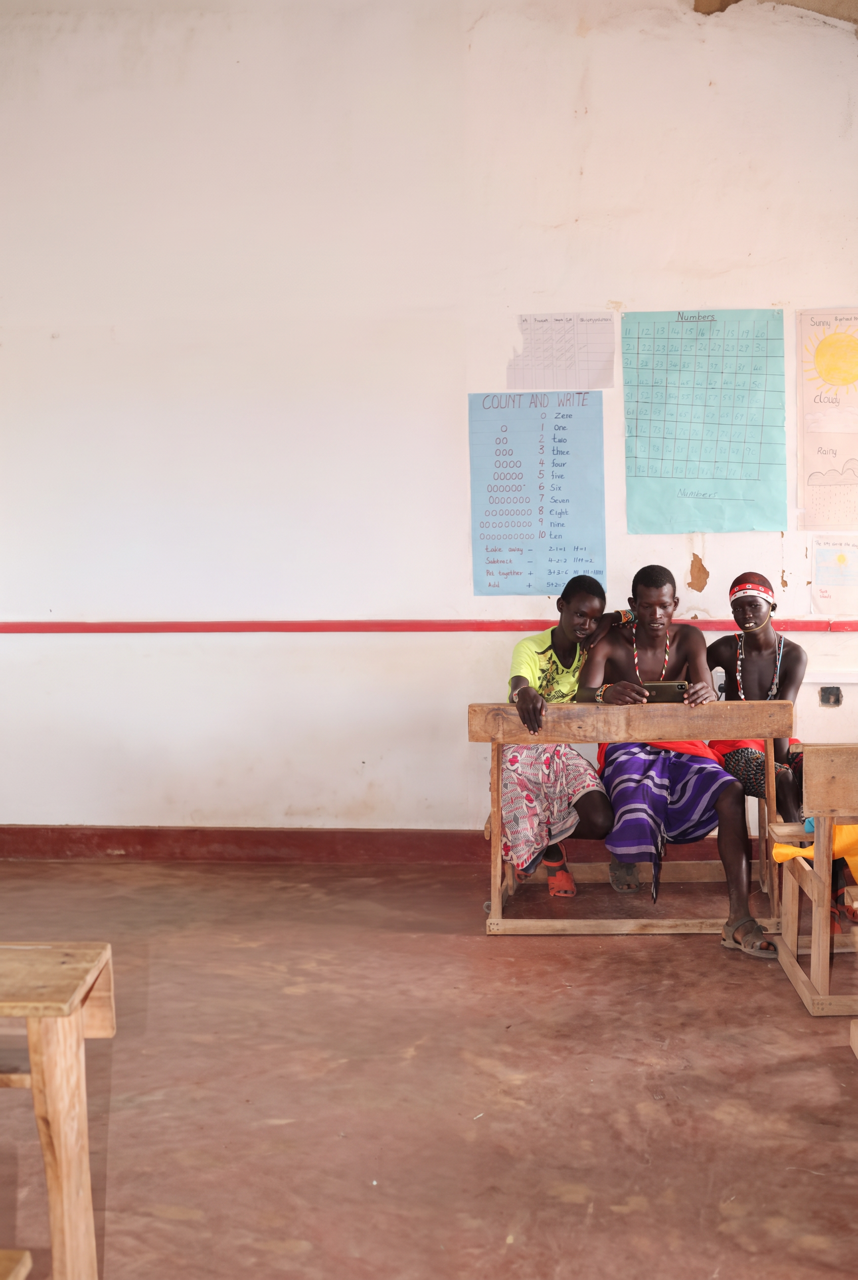 Three young students in a Kenyan classroom sharing a smartphone at a wooden desk